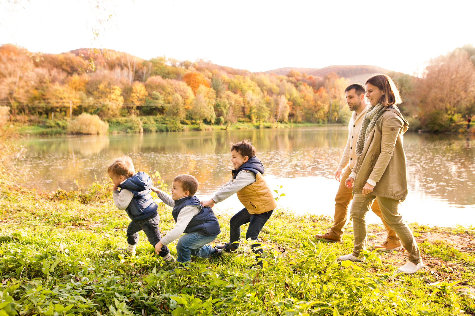 Beautiful young family on a walk at the lake in forest. Mother and father with their three sons in warm clothes outside in colorful autumn nature.