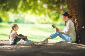 Mother and daughter sitting under the tree on summer lawn. Happy family playing outdoors. Pretty young mom reading a book to her child in the park outside.